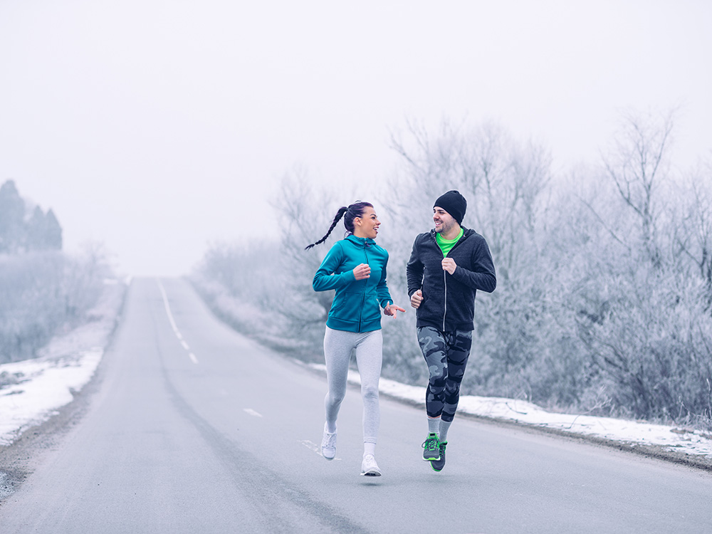 Läuferin und Läufer in Laufbekleidung mit geöffnetem Reisverschluss auf einer Straße im Winter.