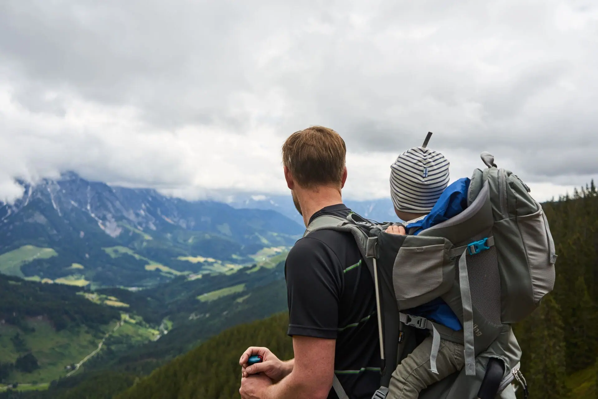 Mann trägt ein Baby in einer Kraxe und schaut auf die Berge.