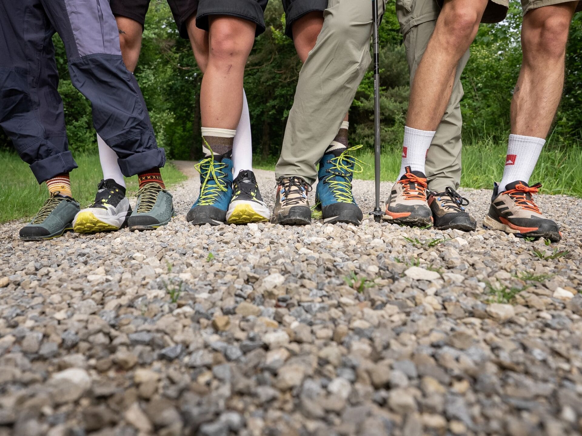 10 Beine mit 5 unterschiedlichen Wanderschuhen an den Füßen stehen ineinander verschränkt auf einem Kiesweg in der Natur.