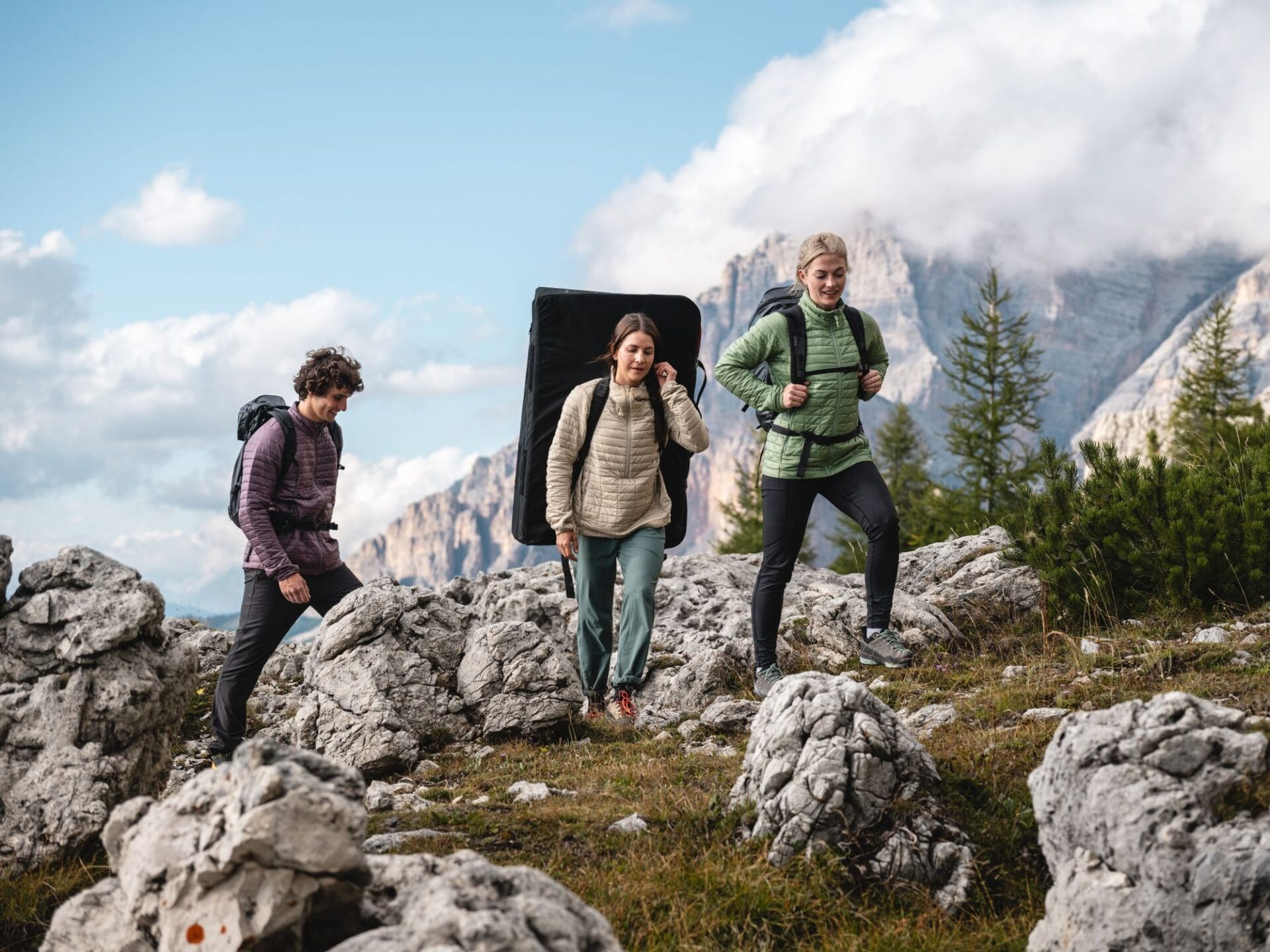 3 Menschen mit Rucksack und Matte auf dem Rücken laufen durch eine alpine Landschaf auf der Suche nach einem passenden Boulderspot.
